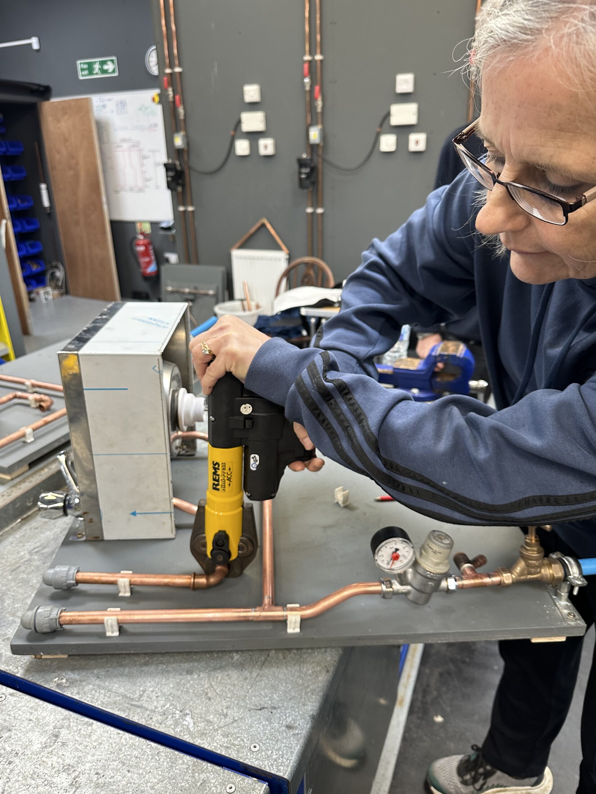 A female is working on a metal apparatus with copper pipes and various valves, using a yellow and black mechanical tool. They are focused on their task, adjusting a component on the setup. The background is a workshop environment with tools and electrical panels on the wall.