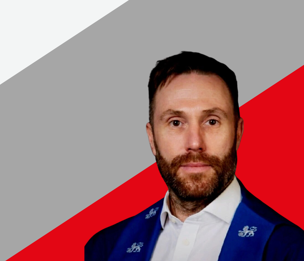 Professional headshot of a man with a beard and short hair wearing a blue blazer with subtle leaf motifs, a white shirt, and a tie. He is superimposed over a geometric background divided diagonally into a grey upper left section with the Belfor Property Restoration logo and a bright red lower right section.