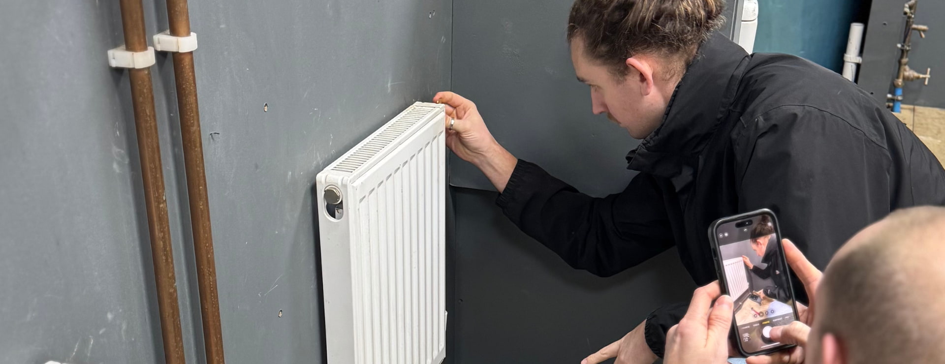 a plumbing student bleeding a radiator as part of their training with his classmate photographing it